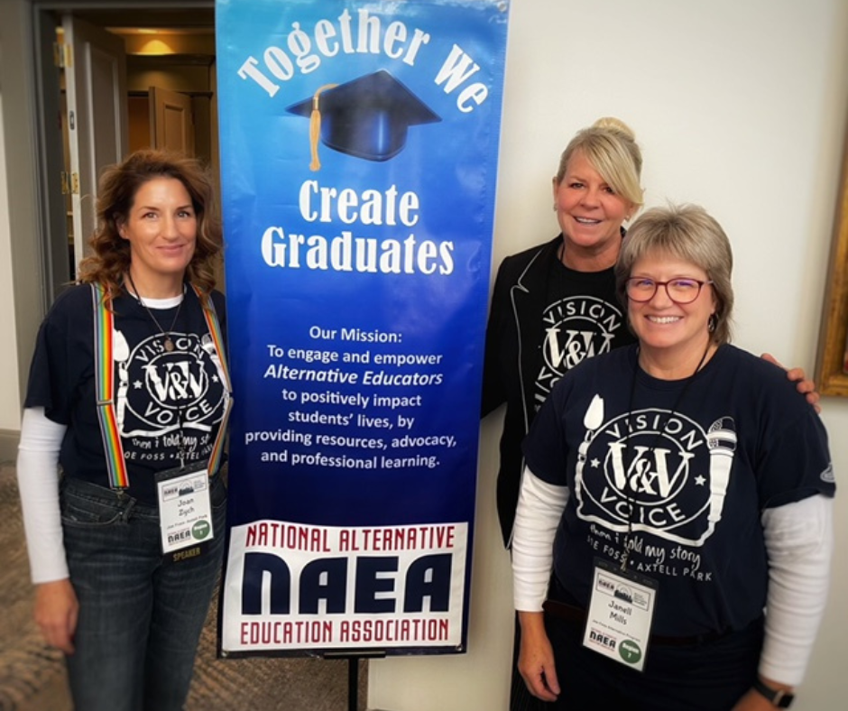Three women dressed in a navy blue tshirt that reads "vision and voice" stand in front of a sign that says "together we create graduates"