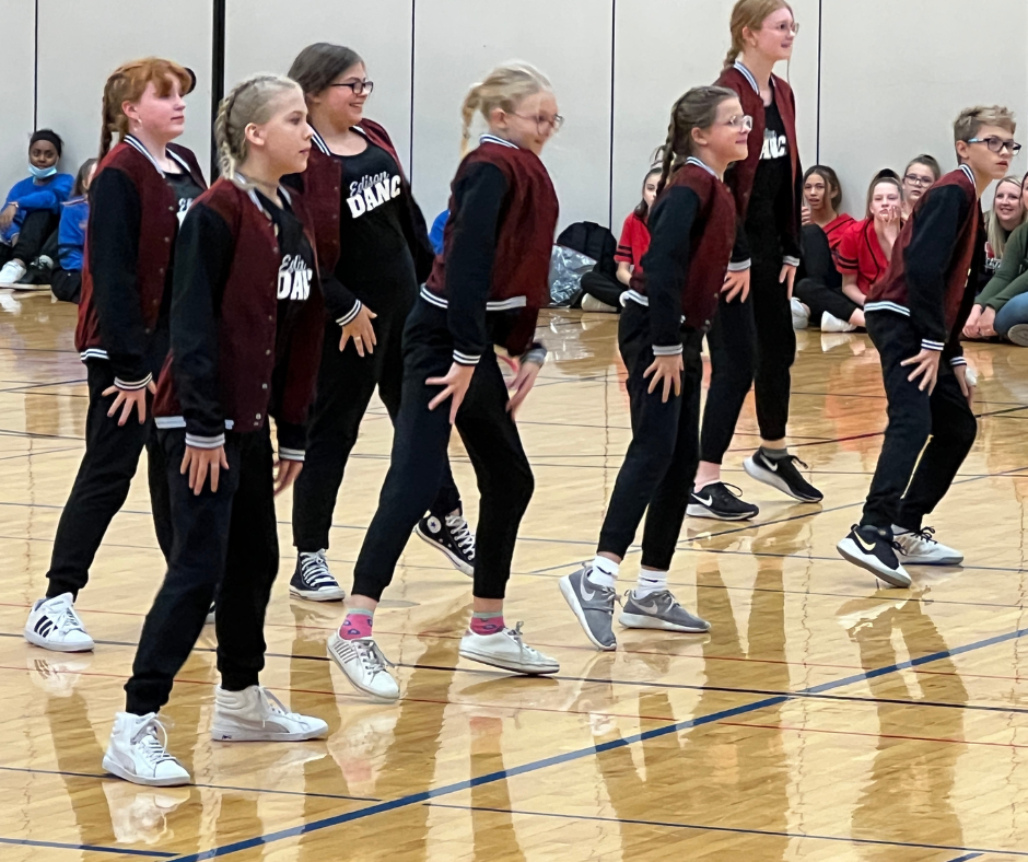 group of 7 middle school students wearing maroon lettermans jacket and black pants with white shows on a gym floor performing a dance routine