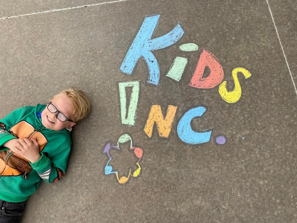 Young boy with pale skin, blonde hair and glasses dressed in a ninja turtles hooded sweatshirt laying on the sidewalk next to a colorful kids inc chalk drawing 